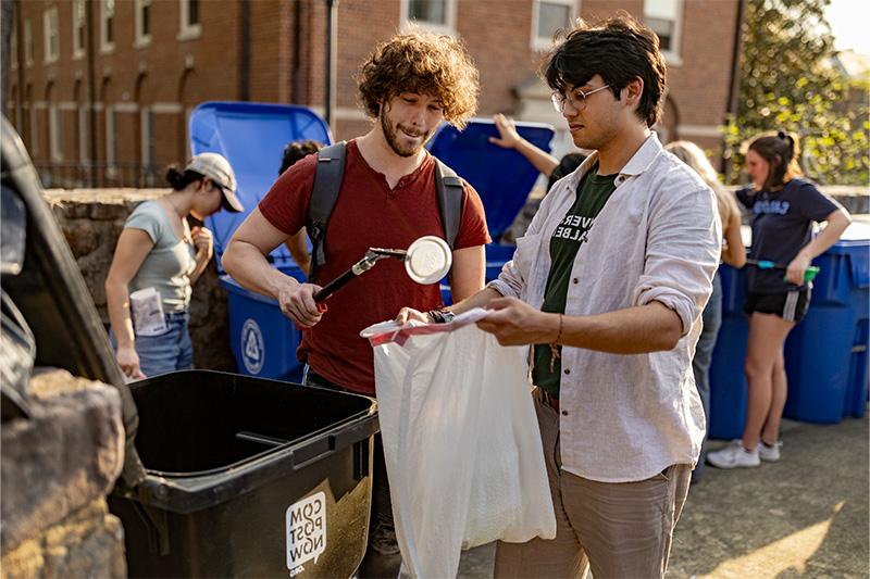 Two students, one holding a trash bag and the other placing a plastic lid inside of it.